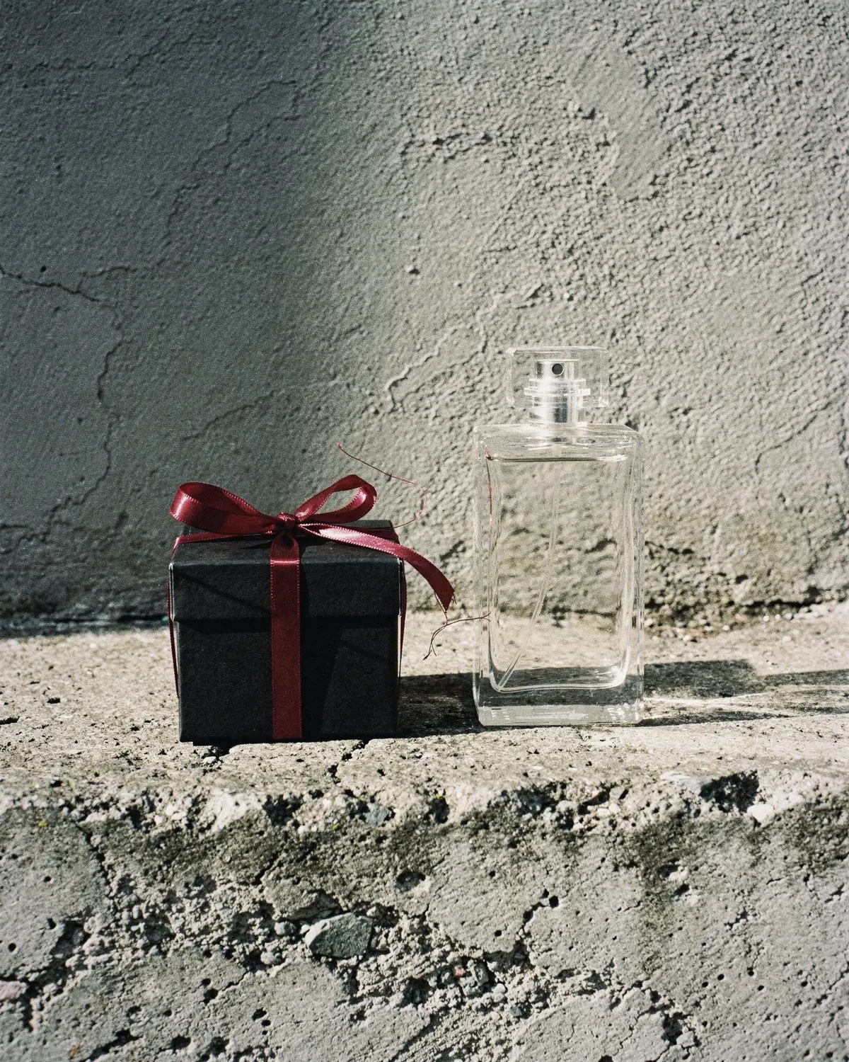 A small black gift box tied with a red satin ribbon beside a clear glass bottle on a concrete ledge in hard winter sun