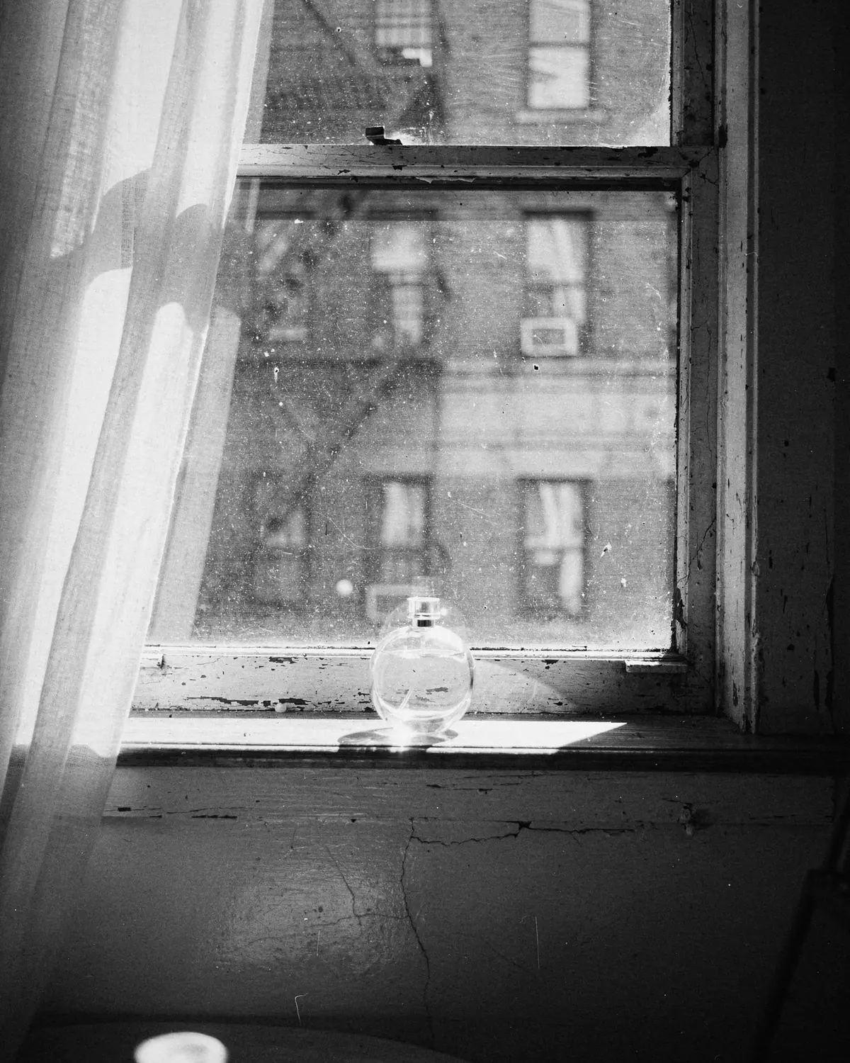 Black and white: a round glass bottle on an old apartment windowsill beside a sheer curtain, fire escapes outside