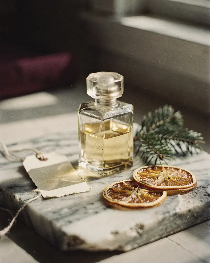 An unlabeled glass bottle on a chipped marble slab with a blank gift tag, a fir sprig and dried orange slices in low winter sun