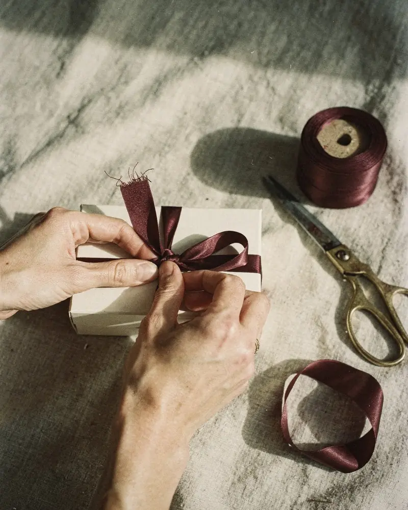 Two hands tying a bordeaux silk ribbon around a small ivory gift box on linen
