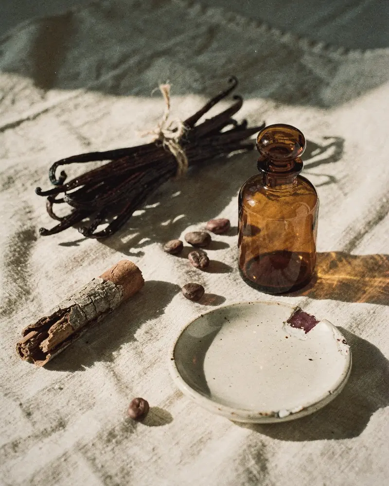 A small amber glass bottle beside vanilla pods, tonka beans and cinnamon bark on crumpled linen